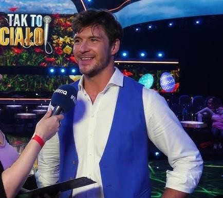 Mikołaj Roznerski na scenie teatralnej podczas występu, oświetlony światłami reflektorów.