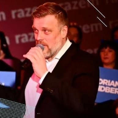 Alt: Adrian Zandberg uśmiechnięty, w luźnej rozmowie z mieszkańcami podczas spotkania publicznego, w tle widać niewielki tłum.