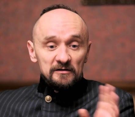 Zamyślony Jan Klata, reżyser teatralny, z intensywnym spojrzeniem, na tle rozmytego światła scenicznego.