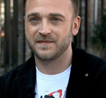 Mateusz Gessler w akcji w kuchni, przygotowujący danie, otoczony świeżymi składnikami, w kitlu szefa kuchni.