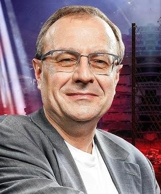 Antoni Dudek podczas wykładu lub wystąpienia telewizyjnego, gestykulujący, z uśmiechem, na tle akademickim.