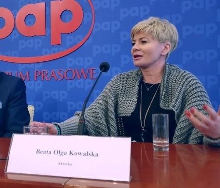 Beata Olga Kowalska na scenie teatralnej, w dynamicznej pozie, pod światłami reflektorów, z tłem sugerującym element scenografii.